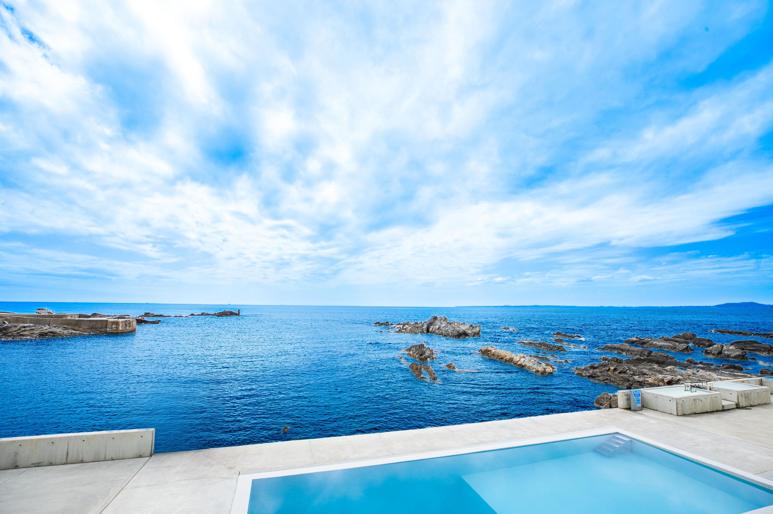 A poolside view overlooking a rocky coastline with clear blue waters and a partly cloudy sky.