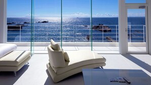 Quarto moderno com uma grande janela de vidro com vista para o mar, com duas poltronas bege e uma pequena mesa com controles remotos.