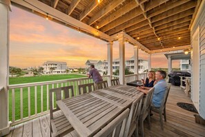 A covered outdoor area with a wooden deck, tables, and chairs, overlooking a grassy area and buildings.