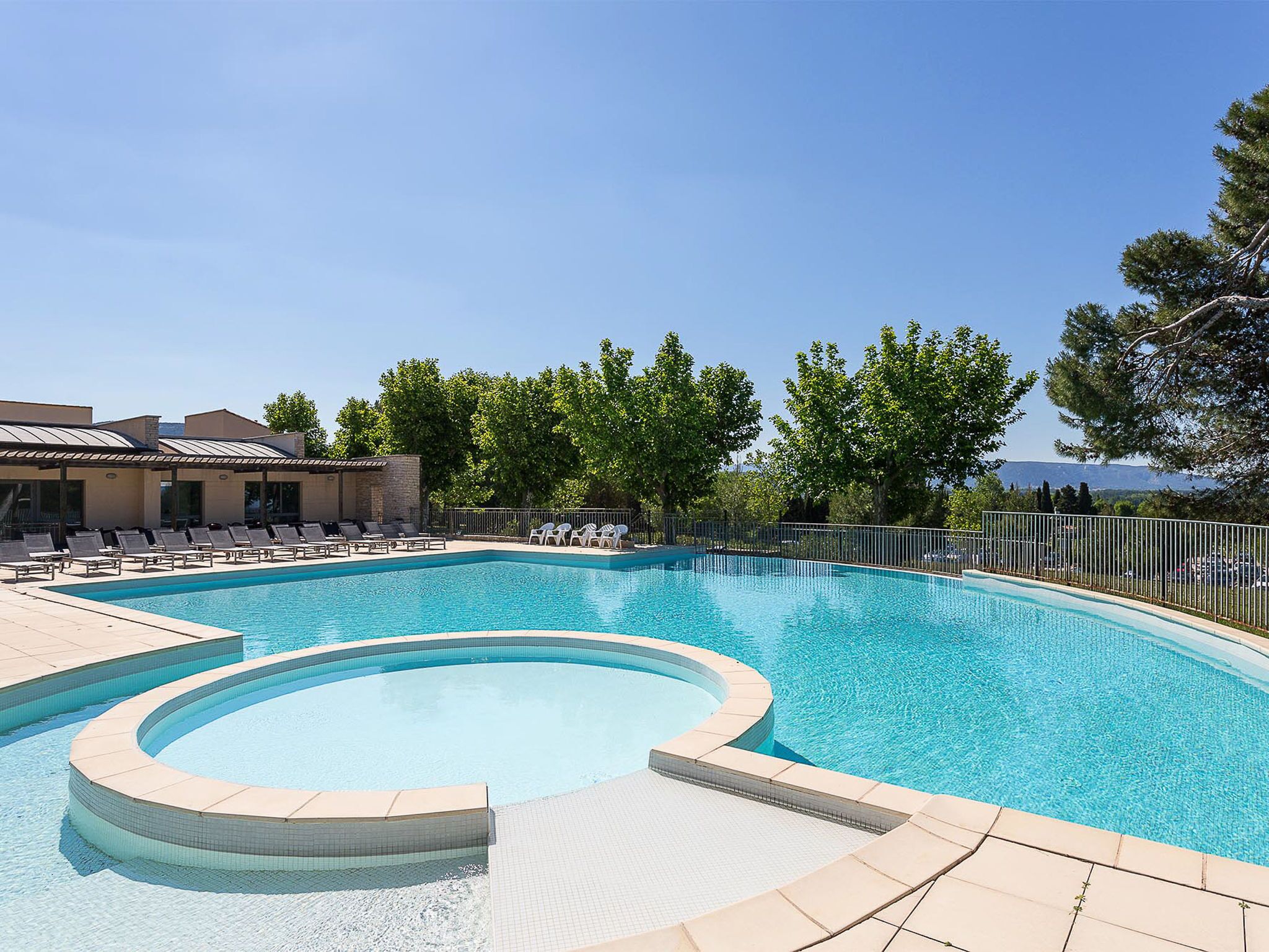 A swimming pool with a curved edge, surrounded by lounge chairs and a building with a tiled roof.