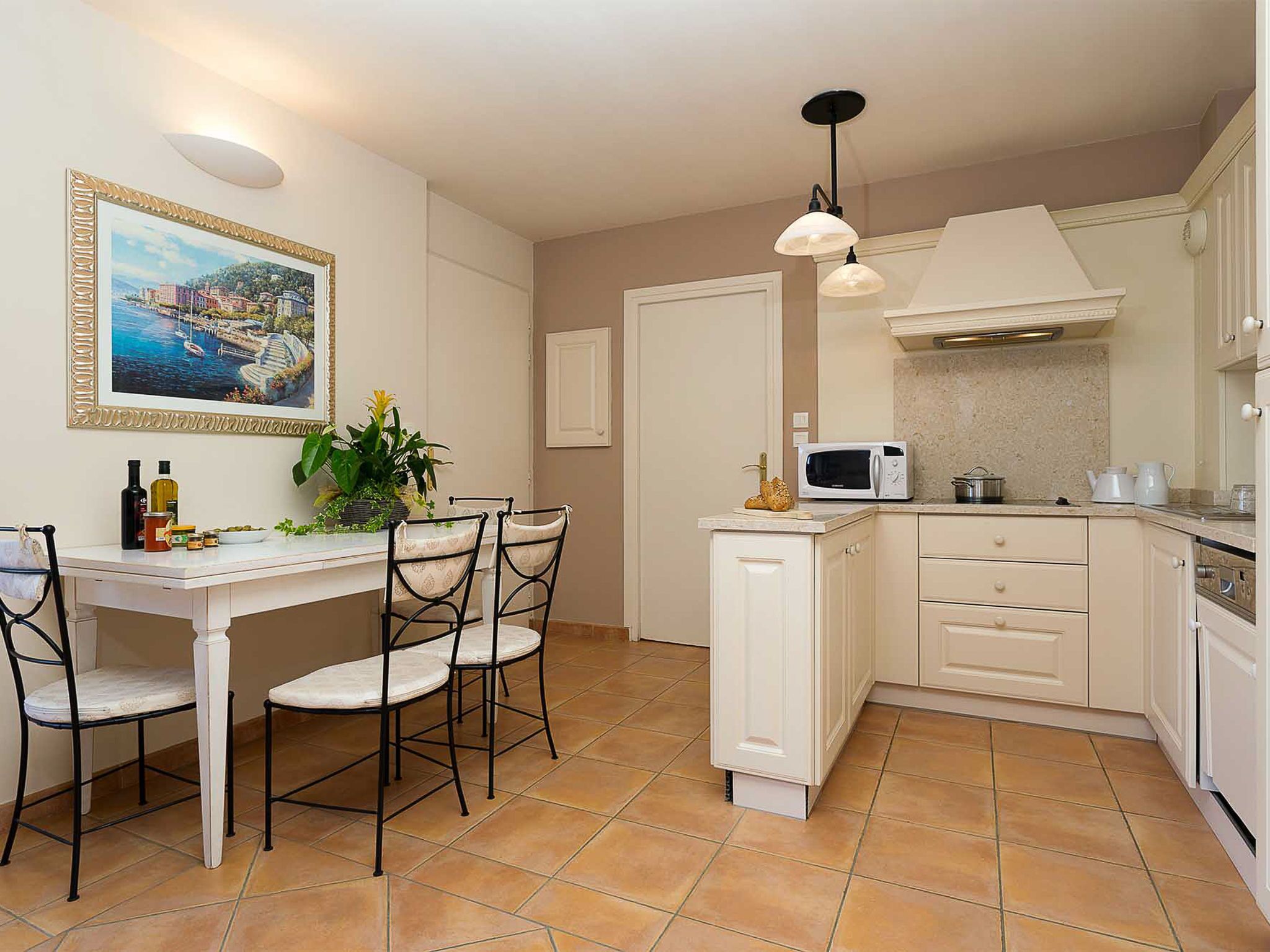 Une cuisine compacte avec des meubles blancs, un four à micro-ondes et un petit îlot central. On y trouve une table à manger avec des chaises et une plante en pot.