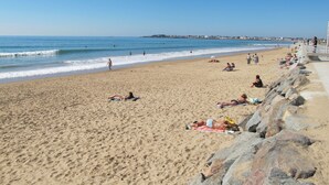 Una playa arenosa con gente tomando el sol y nadando, un rompeolas rocoso y un cielo azul despejado.