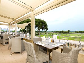 Een buitenterras met rieten stoelen en tafels, een overdekt dak en uitzicht op een golfbaan.