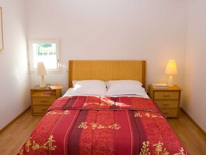 Ein Bett mit einer rot-goldenen Tagesdecke, zwei weiße Kissen und ein Holzkopfstück. Es gibt zwei Nachttische mit Lampen und eine kleine Schublade.