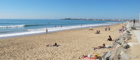 A sandy beach with people sunbathing and swimming, a rocky breakwater, and a clear blue sky.