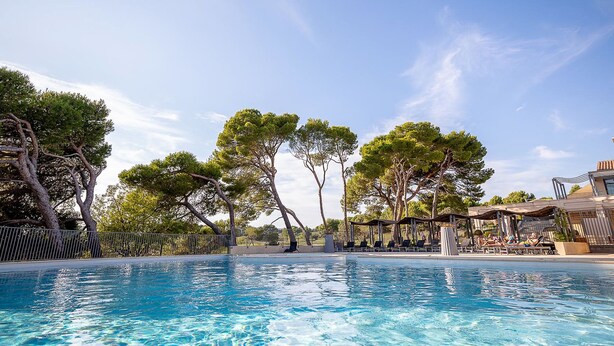 Una piscina con agua cristalina azul, rodeada de árboles y un edificio con mesas al aire libre.