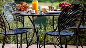 Ein Tisch im Freien mit Frühstück, darunter Croissants, Obst und Saft, mit Stühlen und einer schönen Aussicht im Hintergrund.