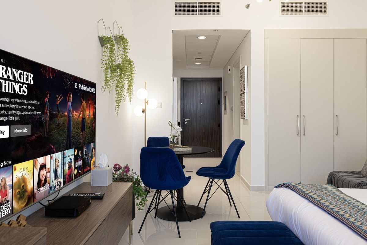 A modern hotel room with a flat-screen TV, a dining area with two chairs, a bed with a patterned bedspread, and a wardrobe.