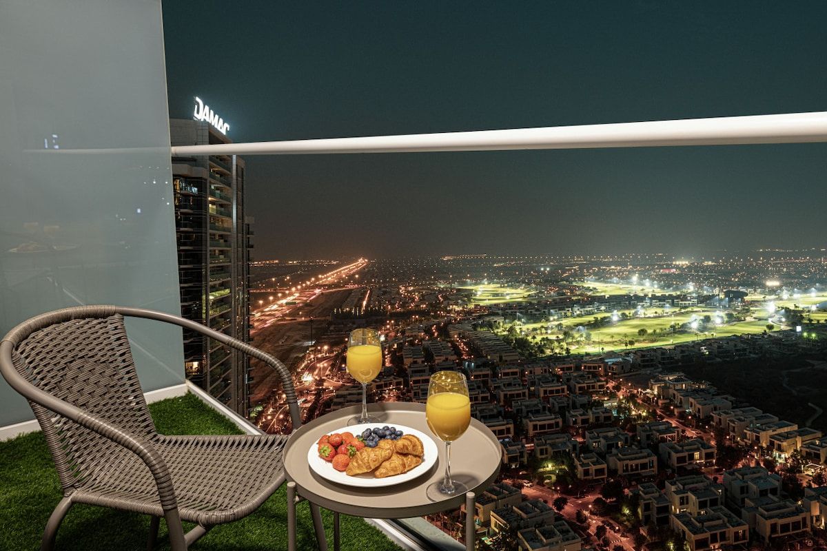 Un balcón con una silla de mimbre, una mesa con comida y bebidas, y vista a la ciudad de noche.