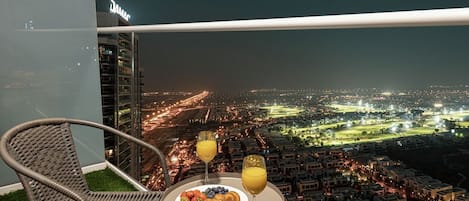 A balcony with a wicker chair, a table with a plate of food and drinks, and a city view at night.