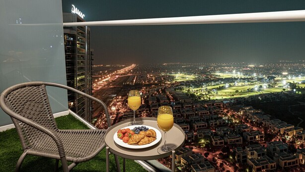 Een balkon met een rieten stoel, een tafeltje met eten en drinken, en een stadsgezicht bij nacht.