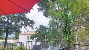Een terras met een tafel en stoelen, een rode parasol en uitzicht op een gebouw en bomen.