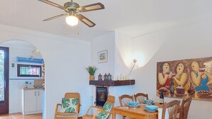 Een eethoek met een houten tafel en stoelen, een plafondventilator, een open haard en een schilderij aan de muur.
