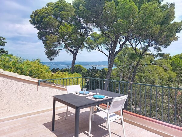A terrace with a table and chairs, overlooking a scenic landscape with trees and a distant view of the sea.