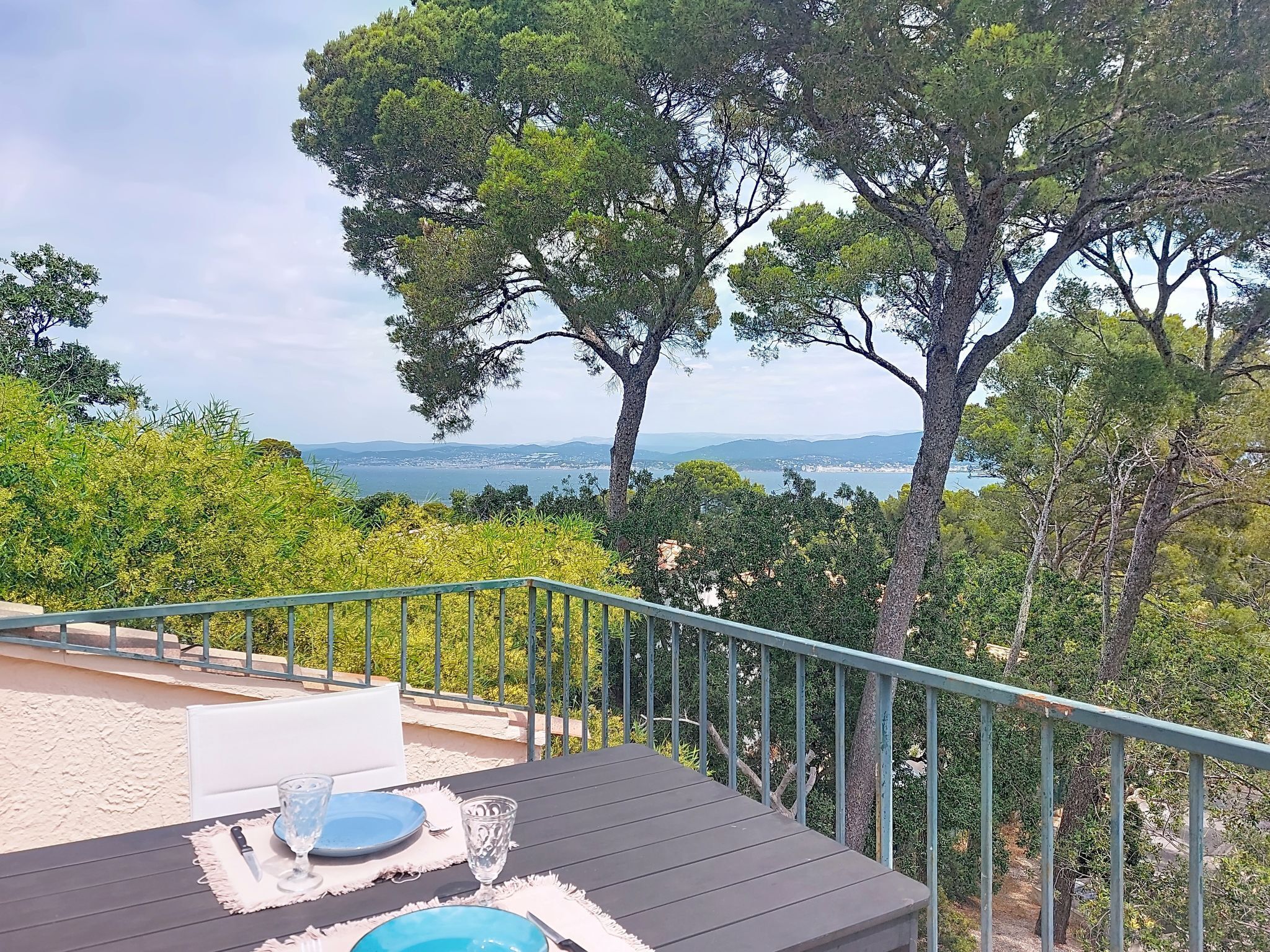 A terrace with a table set for two, overlooking a coastal landscape with trees and a view of the sea.