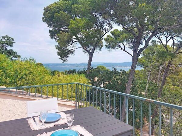A terrace with a table set for two, overlooking a coastal landscape with trees and a view of the sea.