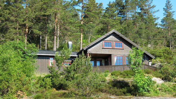 Une cabane avec un toit à pignon, deux fenêtres aux cadres rouges et une terrasse en bois.