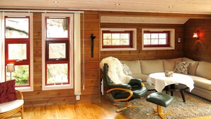 L’intérieur d’un chalet accueillant, avec des murs en bois, un canapé, un fauteuil, une table et de grandes fenêtres dotées de rideaux.