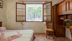 Un dormitorio con un ventanal grande, una cama con ropa de cama floral, una silla de madera y un mueble de madera con libros y flores.