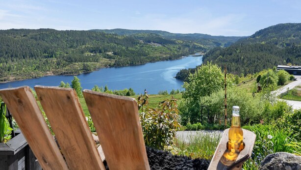 Ein Holzstuhl und eine Bierflasche mit Blick auf einen See, umgeben von bewaldeten Hügeln.