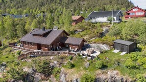 Eine Holzhütte mit einer Terrasse, umgeben von einem bewaldeten Hang und einem Berg im Hintergrund.