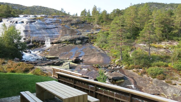Una terrazza in legno con panchine che si affaccia su una cascata rocciosa e colline boscose.