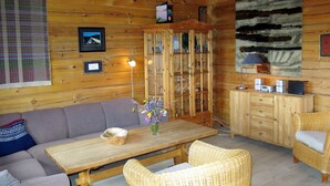 Un accogliente interno di una baita con un tavolo da pranzo in legno, sedie in vimini, un divano, un mobile vetrina con bicchieri e una televisione a parete.