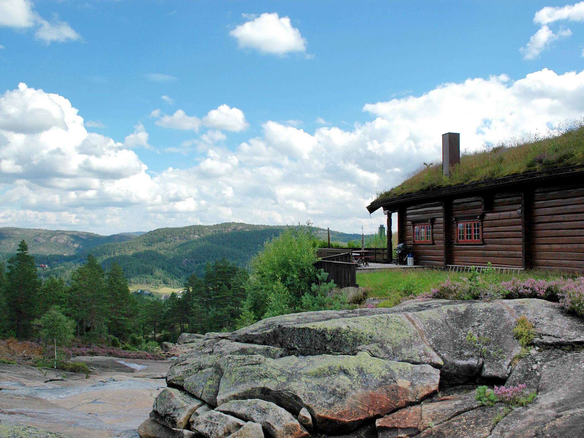 A rustic log cabin with a thatched roof, surrounded by a rocky landscape and a view of distant mountains.