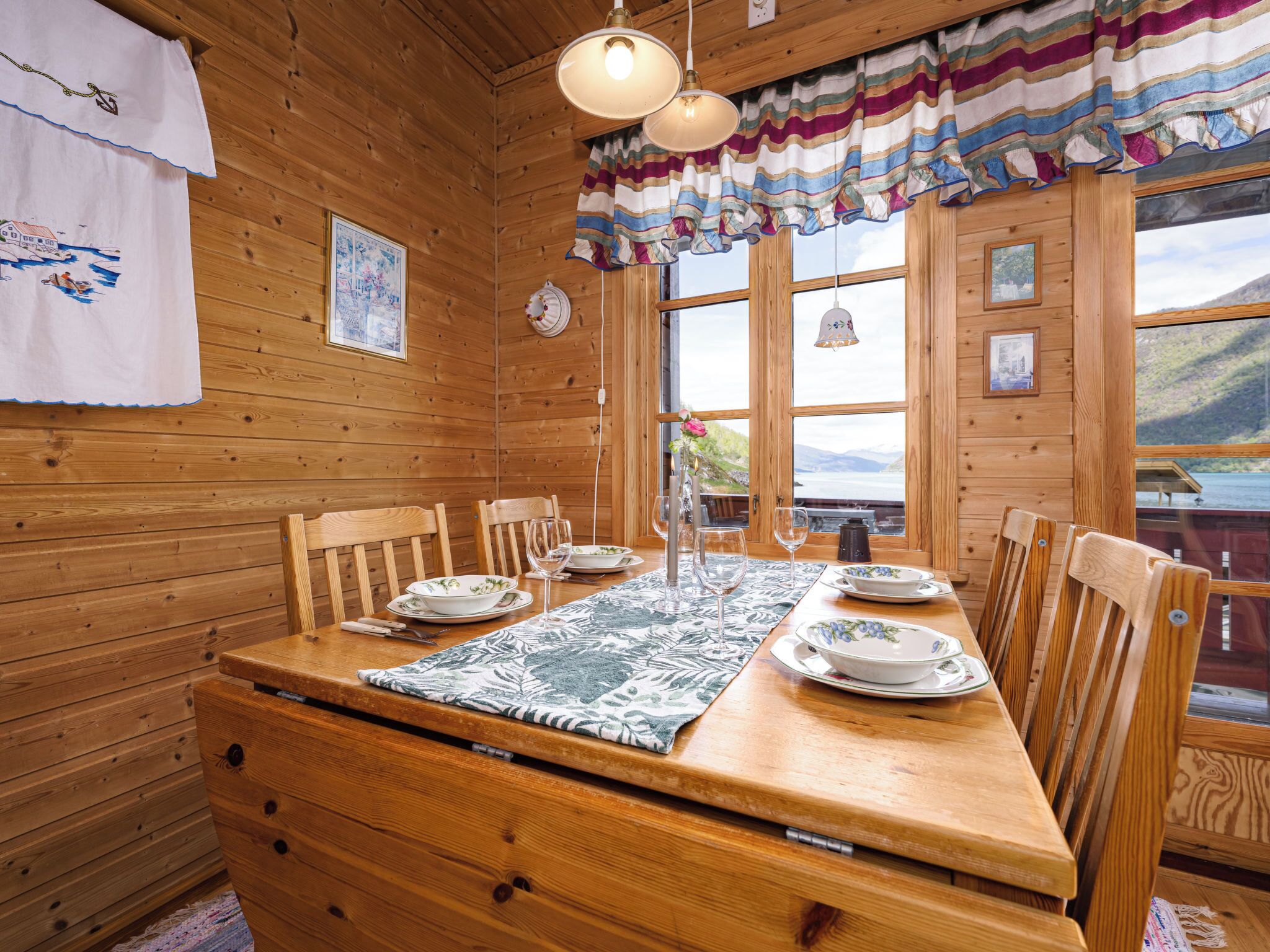 Interior de cabana de madeira, com mesa para quatro pessoas, cadeiras de madeira, um jogo americano estampado e vista do exterior por grandes janelas.