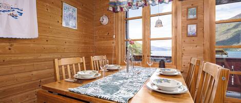 Interior de cabana de madeira, com mesa para quatro pessoas, cadeiras de madeira, um jogo americano estampado e vista do exterior por grandes janelas.