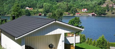 A cabin with a covered porch, a grill, and a view of a lake and forested hills.