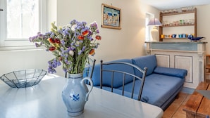 En hyggelig opholdsstue med en blå sofa, et spisebord med en blomstervase, og et køkkenområde med reoler og en lampe.