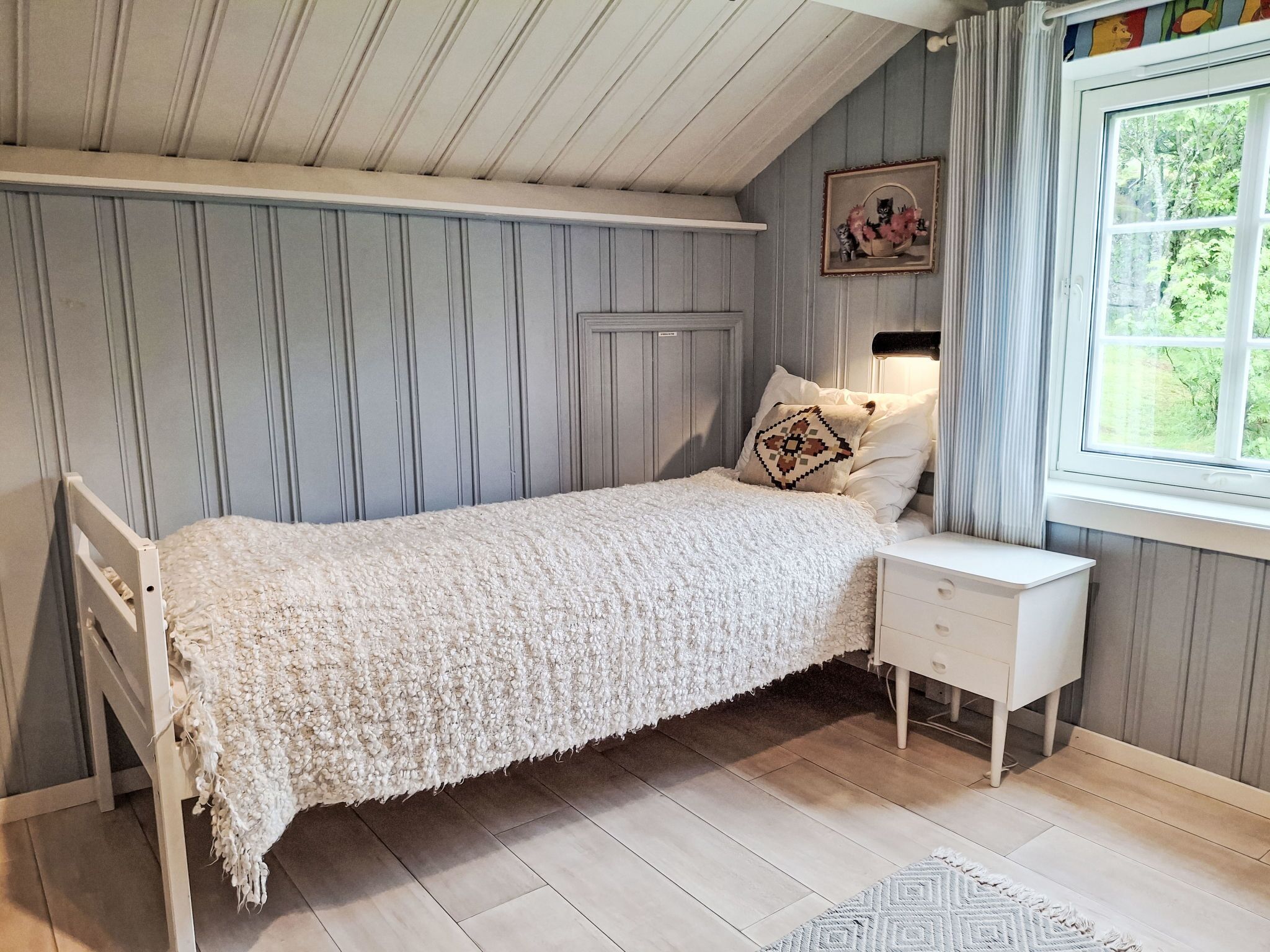 Ein Einzelbett mit weißer Decke, ein Nachttisch, ein Fenster mit Vorhängen und ein Wandbild.