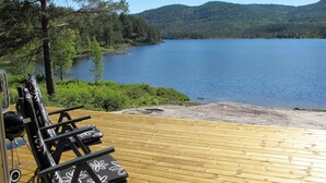 Een houten terras met lounge stoelen, uitkijkend over een meer met een bebost heuvel in de achtergrond.