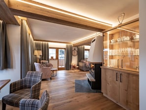 A cozy living room with a fireplace, wooden flooring, and a dining area with glassware. - Luxury apartment close to the ski bus (Itter)