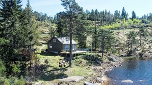 Une cabane au bord d’un lac, dotée d’une terrasse et entourée d’arbres, avec une rive rocheuse.
