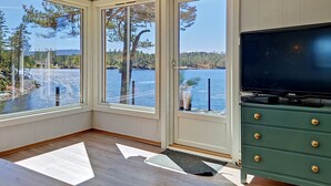 A room with large windows overlooking a lake, a green cabinet with drawers, and a black television. - Vacation home Ogge by Interhome (Vatnestrøm)