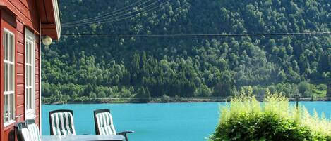 A wooden deck with a table and chairs overlooking a lake, surrounded by lush greenery and a forested hillside.