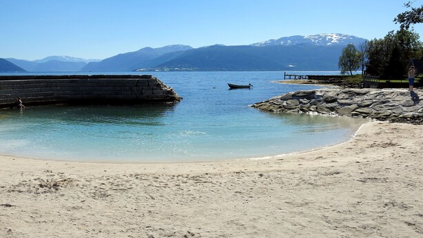 Ein Strand mit klarem Wasser, einem Felsen und Bergen im Hintergrund.