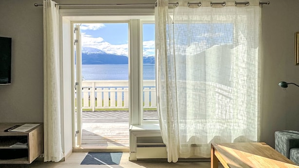 Una habitación con vistas a un lago y montañas, un banco de madera, una mesita y una puerta con cortinas.