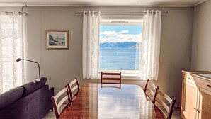 Un comedor con mesa y sillas de madera, un sofá, una lámpara y vista al mar a través de una ventana con cortinas.