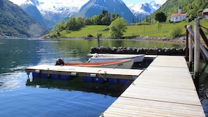 Un muelle de madera se extiende sobre un lago tranquilo, con barcos amarrados al final. Al fondo, montañas nevadas y una casa pequeña con techo rojo.
