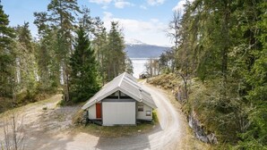 Une cabane avec une allée en gravier, entourée d’une forêt dense et bordée par un lac au loin.
