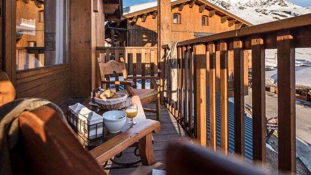 Un balcón de madera con una mesa lista para el desayuno, con vistas a un paisaje montañoso nevado.
