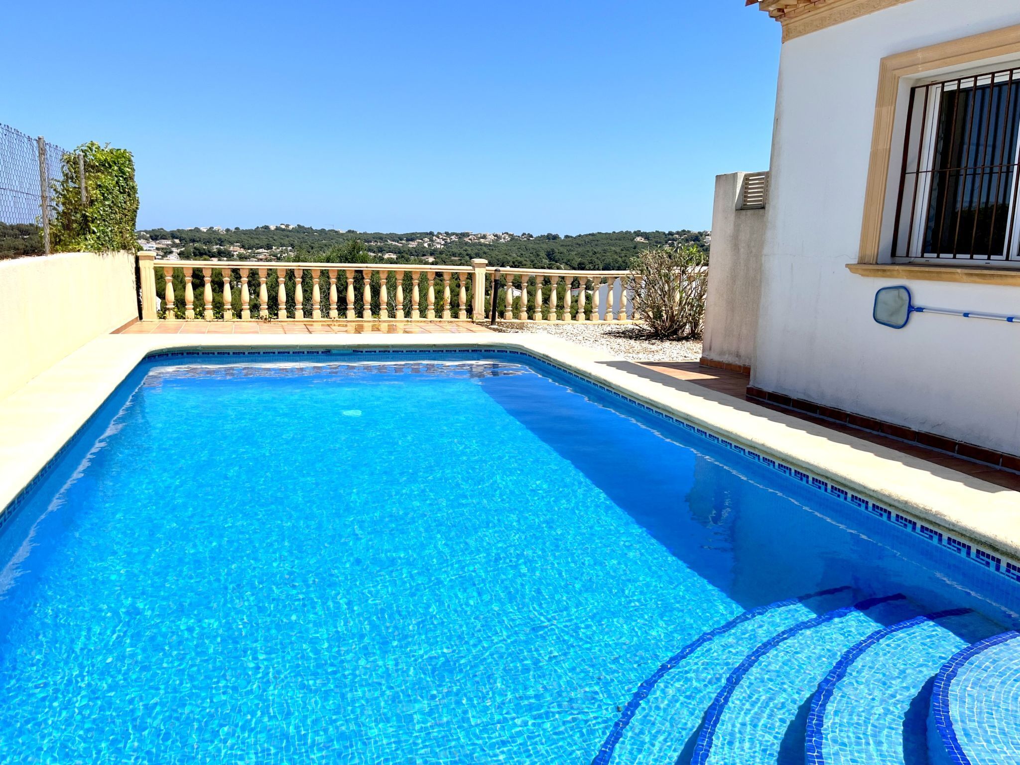 A clear blue swimming pool with a tiled edge, a balcony with a railing, and a view of a hillside with buildings.