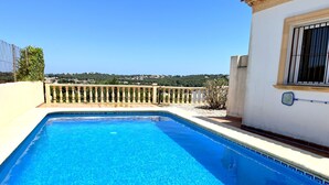 Une piscine d’un bleu éclatant, bordée de carreaux, un balcon avec une balustrade et une vue sur une colline parsemée de bâtiments.
