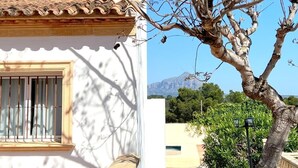 Un bâtiment blanc avec un toit recouvert de tuiles, une fenêtre ornée de barreaux verticaux et un arbre aux branches nues.