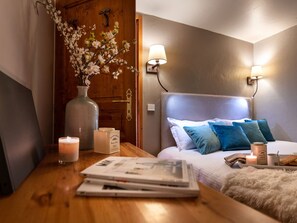 Een slaapkamer met een houten deur, een nachtkastje met een kaars, een lamp en een bed met blauwe kussens.