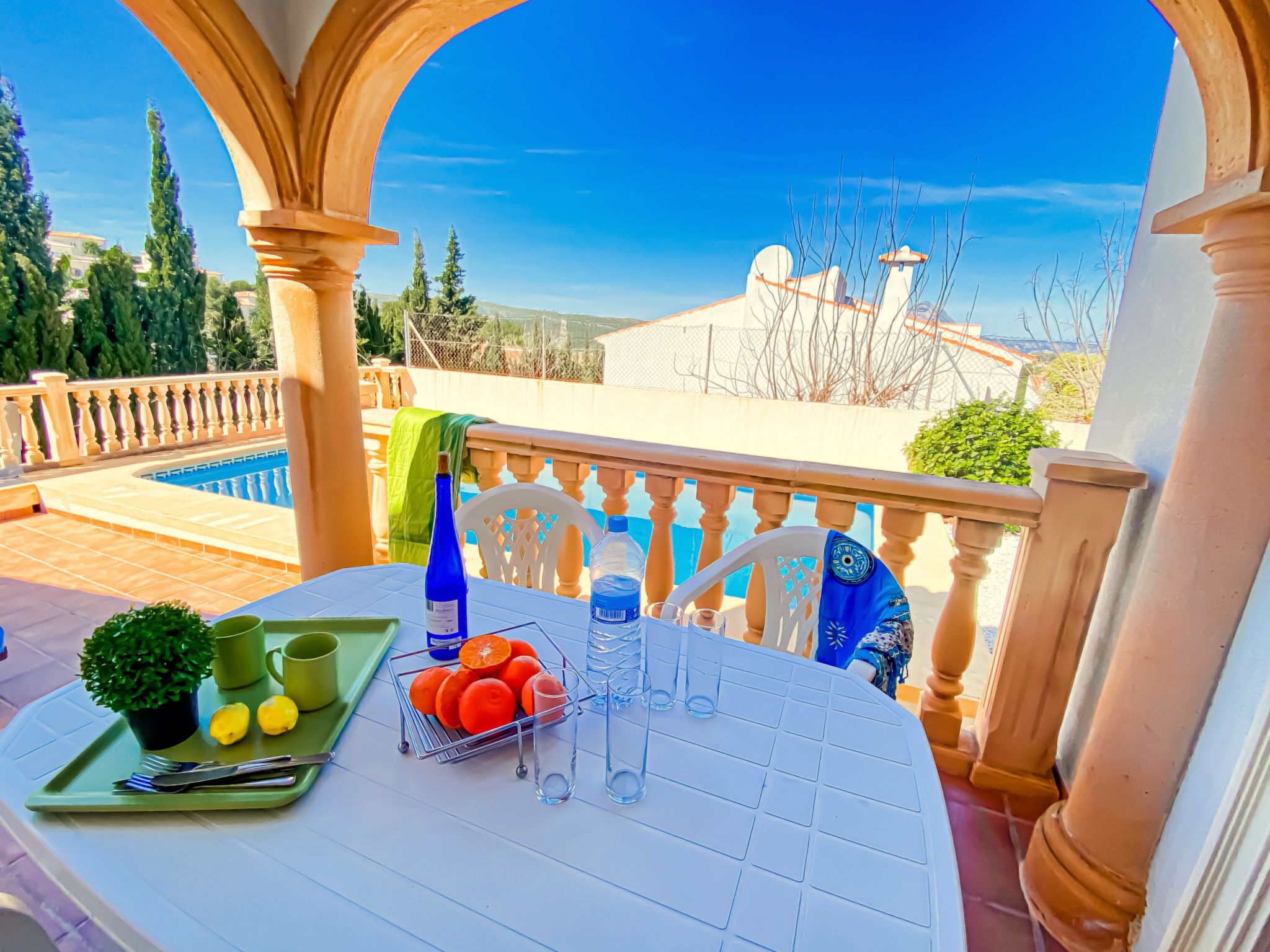 A table set with a green tray, a potted plant, lemons, oranges, and a bottle of water on a balcony overlooking a pool and a clear sky.