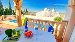 Une table dressée avec un plateau vert, une plante en pot, des citrons, des oranges et une bouteille d’eau, sur un balcon donnant sur une piscine et un ciel dégagé.